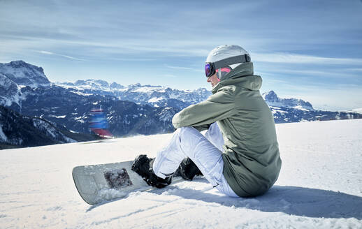 Smiling woman sitting with snowboard on viewpoint and enjoying view at distance - DIKF00391