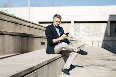 Grauhaariger Geschäftsmann sitzt auf einer Treppe und benutzt ein Mobiltelefon - JRFF04185