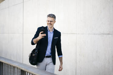 Smiling gray-haired businessman walking in the city using cell phone - JRFF04192