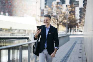 Lächelnder grauhaariger Geschäftsmann, der in der Stadt spazieren geht und ein Handy benutzt - JRFF04193