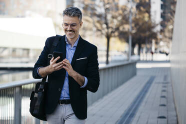 Smiling gray-haired businessman walking in the city using cell phone - JRFF04194