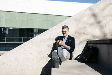Grauhaariger Geschäftsmann sitzt auf einer Treppe und benutzt ein Mobiltelefon - JRFF04200