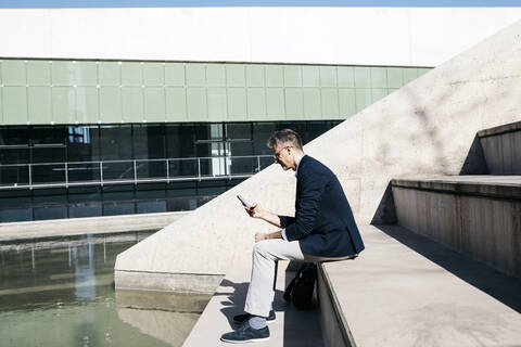 Grauhaariger Geschäftsmann sitzt auf einer Treppe an einem Pool und benutzt ein Mobiltelefon, lizenzfreies Stockfoto