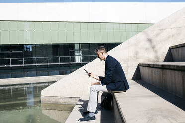 Gray-haired businessman sitting on stairs at a pool using cell phone - JRFF04202