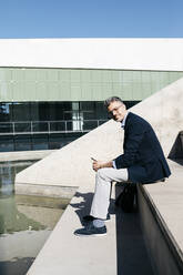 Gray-haired businessman sitting on stairs at a pool - JRFF04203