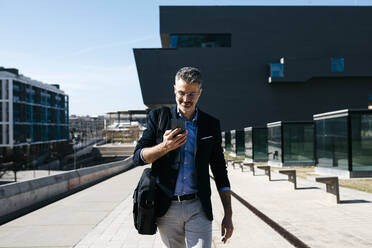 Gray-haired businessman walking with cell phone in the city - JRFF04213