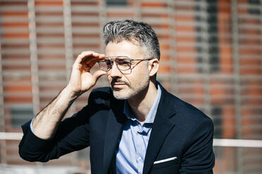 Portrait of a gray-haired businessman in the city putting on glasses - JRFF04217