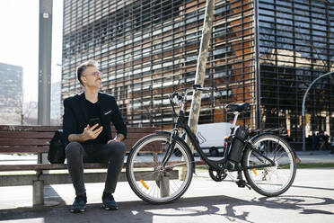 Gray-haired businessman sitting on a bench next to electric bicycle in the city - JRFF04227