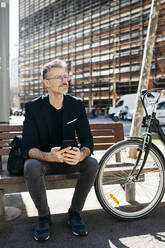 Gray-haired businessman sitting on a bench next to bicycle in the city - JRFF04228