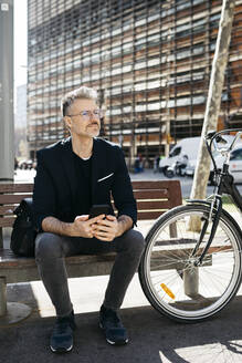 Grauhaariger Geschäftsmann sitzt auf einer Bank neben einem Fahrrad in der Stadt - JRFF04228