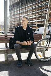 Grauhaariger Geschäftsmann sitzt auf einer Bank neben einem Fahrrad in der Stadt - JRFF04229