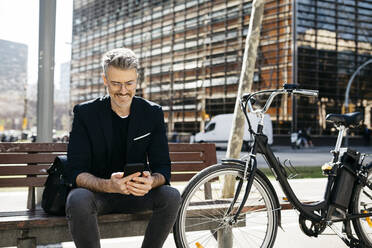 Grauhaariger Geschäftsmann sitzt auf einer Bank neben einem Fahrrad in der Stadt und benutzt ein Mobiltelefon - JRFF04230