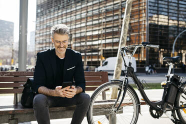 Gray-haired businessman sitting on a bench next to bicycle in the city using cell phone - JRFF04230