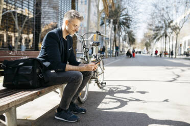 Gray-haired businessman sitting on a bench next to bicycle in the city using cell phone - JRFF04231