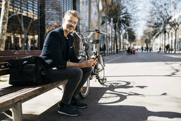 Portrait of gray-haired businessman sitting on a bench next to bicycle in the city - JRFF04232