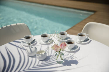 Kaffeetassen und Rosen auf einem sonnigen Terrassentisch im Sommer am Pool - HOXF05327