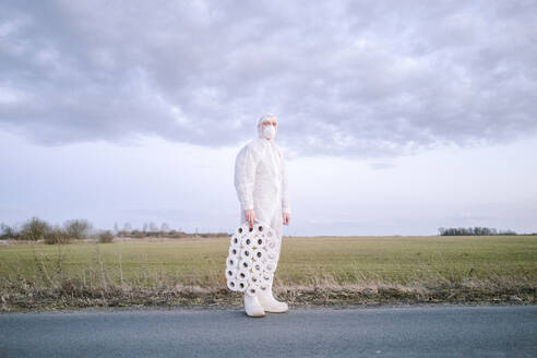 Man wearing protective suit and mask standing on country road with toilet rolls - EYAF00957
