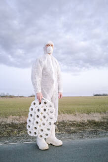 Mann mit Schutzanzug und Maske steht auf einer Landstraße mit Toilettenrollen - EYAF00958