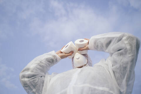 Mann mit Schutzanzug und Maske benutzt Toilettenpapier wie ein Fernglas, lizenzfreies Stockfoto