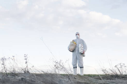 Mann mit Schutzanzug und Maske hält Klopapierrollen und Einkaufstasche in der Landschaft - EYAF00962