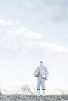 Mann mit Schutzanzug und Maske hält Klopapierrollen und Einkaufstasche in der Landschaft - EYAF00963