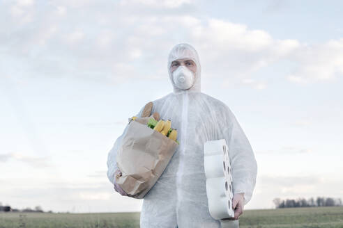 Mann mit Schutzanzug und Maske hält Klopapierrollen und Einkaufstasche in der Landschaft - EYAF00964