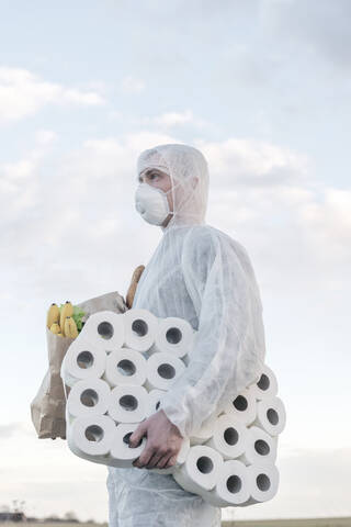 Mann mit Schutzanzug und Maske hält Toilettenpapier und Einkaufstasche im Freien, lizenzfreies Stockfoto