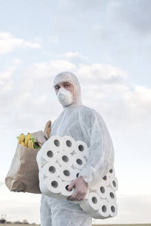 Mann mit Schutzanzug und Maske hält Toilettenpapier und Einkaufstasche im Freien - EYAF00966