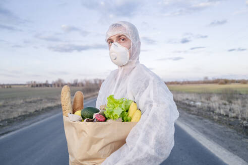 Man wearing protective suit and mask holding grocery bag on a country road - EYAF00969