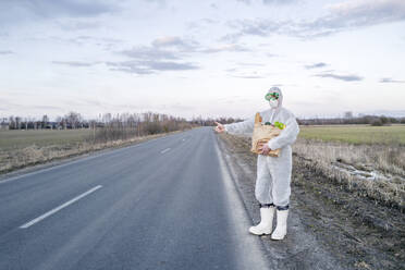 Mann mit Schutzanzug und Maske, der eine Einkaufstasche in der Hand hält und auf einer Landstraße trampen geht - EYAF00973