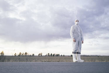 Man wearing protective suit and mask at the roadside of a country road - EYAF00980