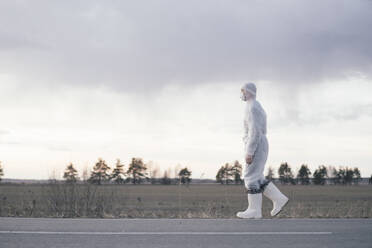 Man wearing protective suit and mask at the roadside of a country road - EYAF00981