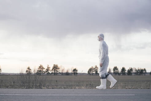 Man wearing protective suit and mask at the roadside of a country road - EYAF00981
