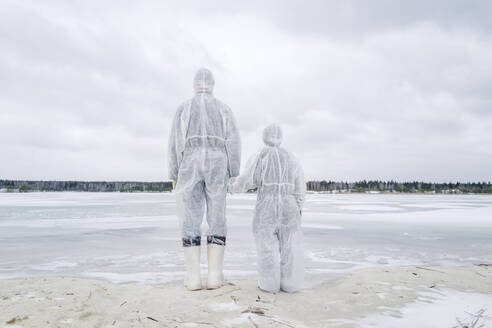 Rückansicht von Vater und Sohn in Schutzanzügen, die am zugefrorenen Fluss stehen - EYAF00982