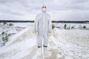 Man wearing protective suit and mask standing in rural winter landscape - EYAF00991