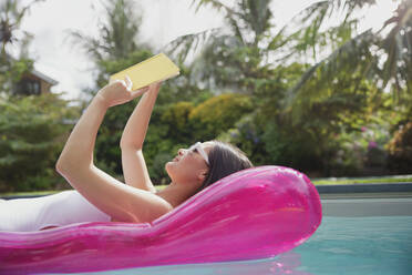 Frau liest Buch auf aufblasbarem Floß in sonnigem Sommerschwimmbad - HOXF05451