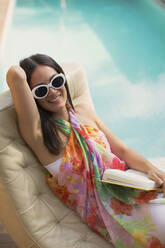 Portrait happy woman relaxing, reading book on lounge chair at summer poolside - HOXF05461