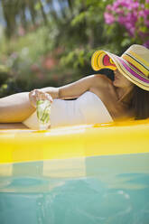 Sinnliche Frau im Badeanzug und Sonnenhut entspannt sich mit einem Cocktail auf einem aufblasbaren Floß im sonnigen Schwimmbad - HOXF05469