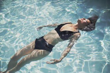 Serene, sensual woman in black bikini relaxing, floating in sunny summer swimming pool - HOXF05470