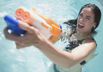 Porträt verspielte Frau mit Spritzpistole in sonnigem Schwimmbad - HOXF05475