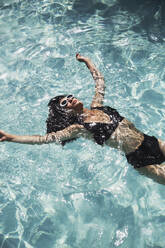Serene woman in black bikini floating in sunny summer swimming pool - HOXF05478