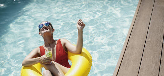 Unbeschwerte Frau mit aufblasbarem Ring trinkt Cocktail im sonnigen Schwimmbad - HOXF05491