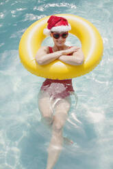 Portrait smiling woman in Santa hat and heart-shape sunglasses swimming with inflatable ring in swimming pool - HOXF05496