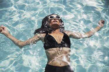 Serene woman in black bikini floating in sunny summer swimming pool - HOXF05503