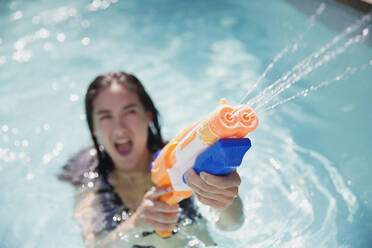 Playful woman using squirt gun in sunny summer swimming pool - HOXF05504