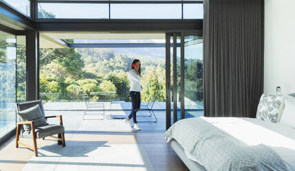 Woman talking on smart phone in patio doorway of modern, luxury bedroom - HOXF05559