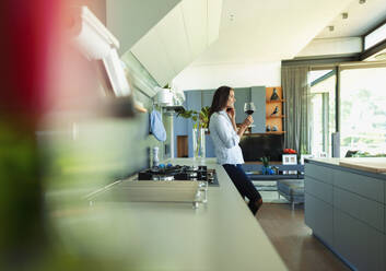 Woman drinking red wine and talking on smart phone in modern kitchen - HOXF05572