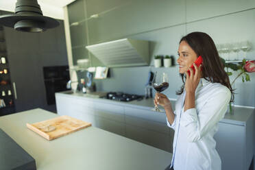 Woman talking on smart phone and drinking red wine in modern kitchen - HOXF05579