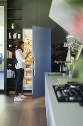 Woman standing at open refrigerator in kitchen - HOXF05585