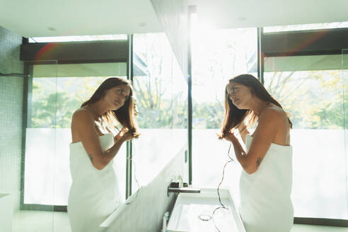 Frau, eingewickelt in ein Handtuch, föhnt sich die Haare im sonnigen Badezimmerspiegel - HOXF05613